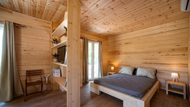Een slaapkamer in een houten hutje, met een bed, nachtkastjes, een stoel en een plank met planten.