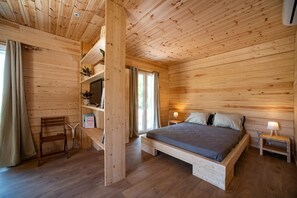 Ett sovrum i en stuga med träpanel, inrett med en säng, två sängbord, en stol och en hylla med växter.