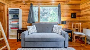 Interior acogedor de una cabaña con un sofá gris, una estantería de madera y una ventana con cortinas.