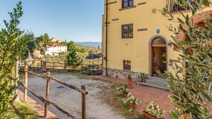 A yellow building with a balcony, a gravel courtyard, and a view of a hill with houses. - Studio 'Il Poderaccio Tramontana' with Shared Pool, Wi-Fi and Air Conditioning (Scandicci)