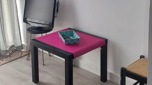 Une pièce avec un téléviseur posé sur un meuble, une table recouverte d’une nappe rose et deux chaises.
