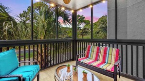 A covered outdoor seating area with wicker furniture, a ceiling fan, and a view of greenery and trees. - Ocean Greens Unit E201 (North Myrtle Beach)