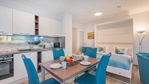 A compact living space with a kitchenette, a dining area with blue chairs, and a bed with a headboard. - Beautiful vacation apartment on Usedom, with terrace and small sunbathing lawn (Zirchow)