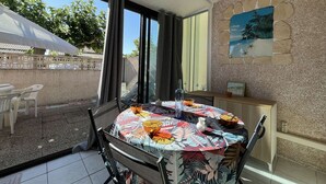 A dining area with a round table covered by a colorful tablecloth, chairs, a window with curtains, and a stone wall with a painting. - Pavilion with Sunny Terrace, Parking - Gruissan (Gruissan)