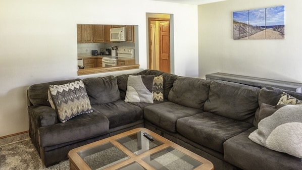 Una sala de estar con un sofá modular, una mesa de centro de vidrio y una zona de cocina con armarios de madera y un microondas.