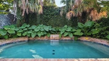 Piscina com borda de tijolos, rodeada por vegetação exuberante e um muro de tijolos.