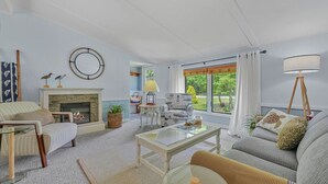 A living room with a fireplace, a sofa, a coffee table, and a view of the outdoors. - Sunny 3BR w/ Firepit + Game Yard | Walk to Green Lake (Interlochen)