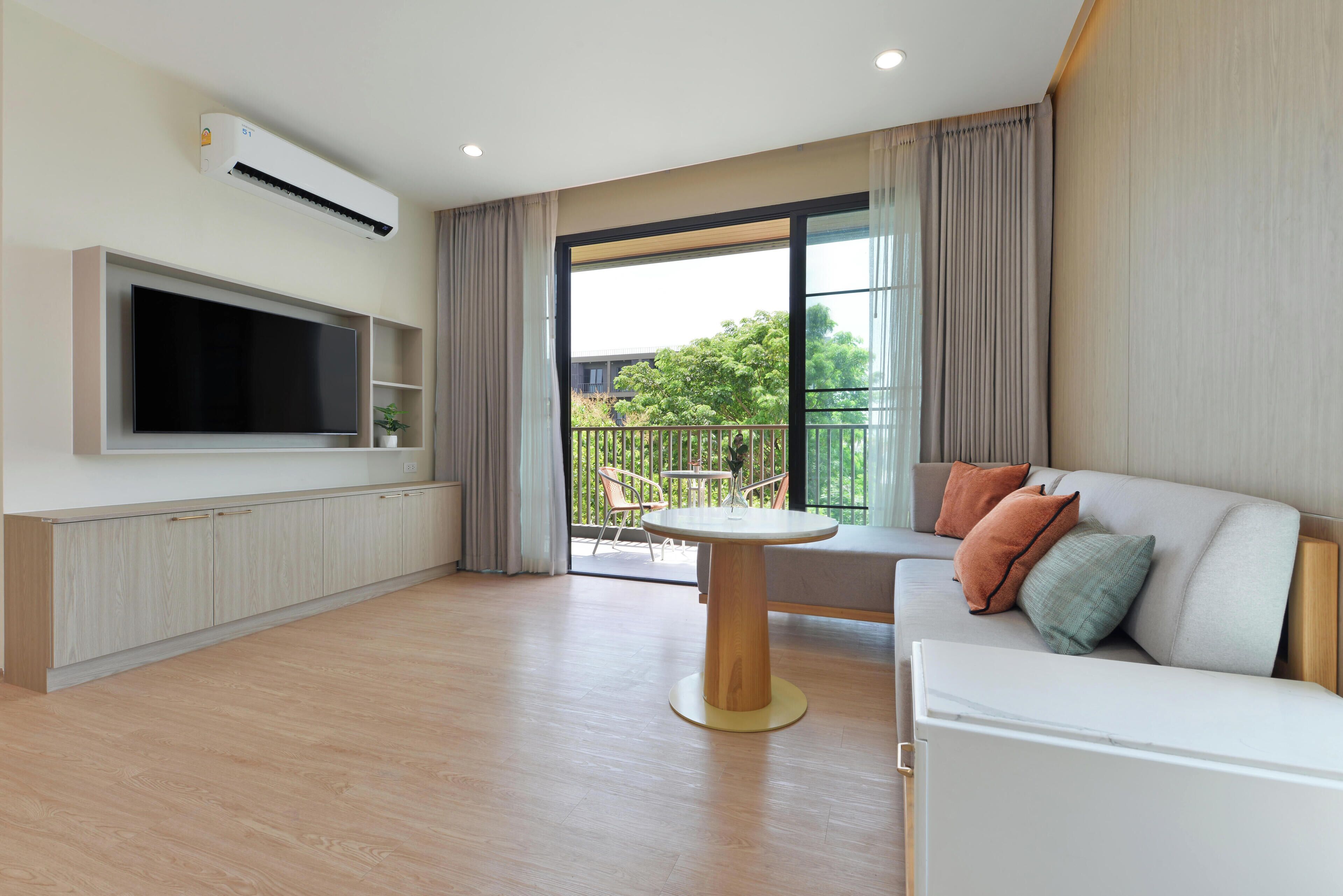 A modern living room with a flat-screen TV, a round wooden table, a sofa with cushions, and a sliding glass door leading to a balcony.
