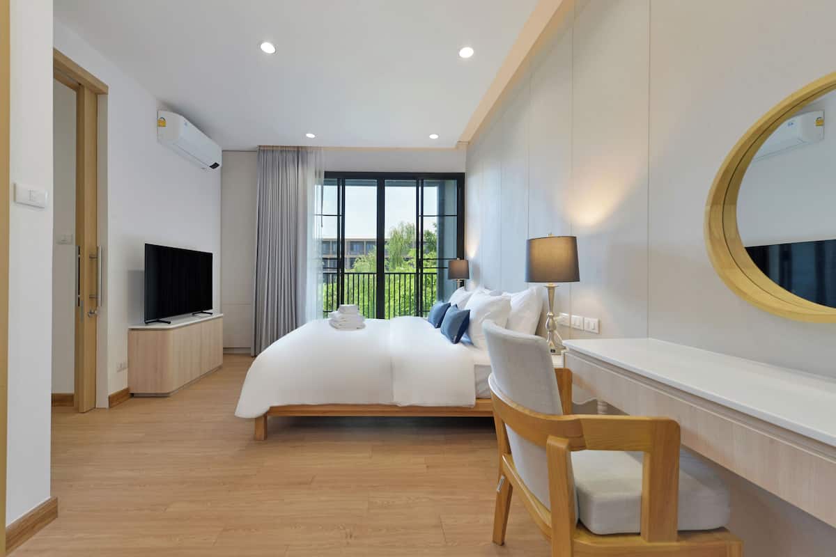 A modern hotel room with a large bed, a TV, a desk with a lamp, and a large window with a view of greenery.