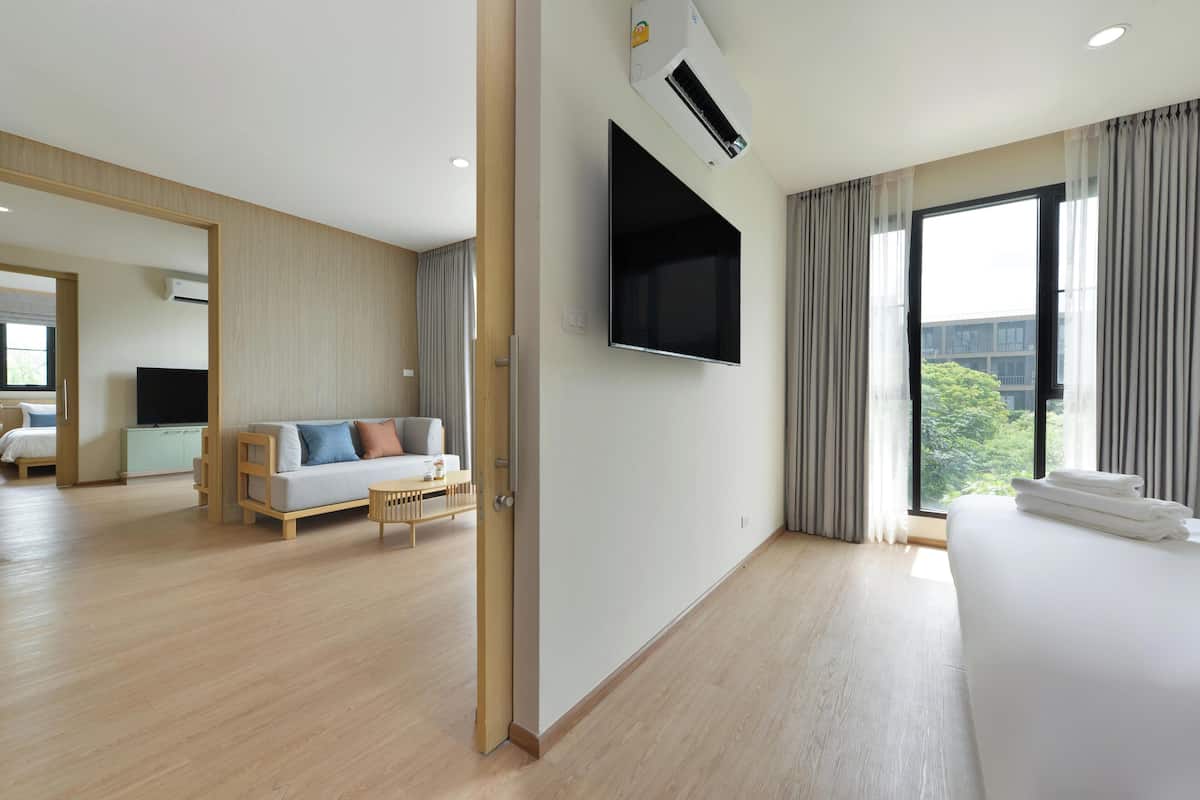 A modern hotel room with a large bed, a flat-screen TV mounted on the wall, a sofa, and a large window with curtains.