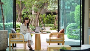 Donna al lavoro su un portatile in una stanza moderna e luminosa, con ampie finestre, mobili in legno e vista su un giardino rigoglioso.