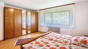 A bedroom with a bed, a wooden wardrobe, a radiator, and a window with sheer curtains.