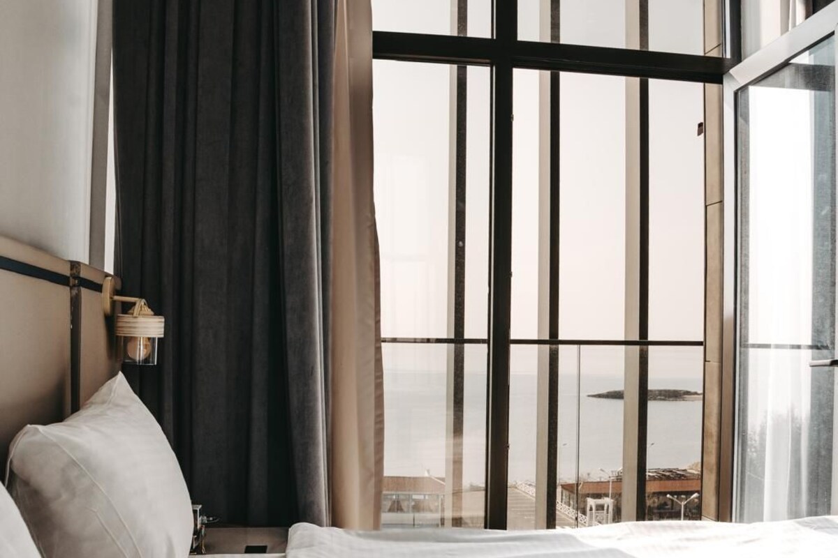 Une chambre d’hôtel avec une grande fenêtre, un lit avec du linge de lit blanc et une table de chevet avec une lampe.