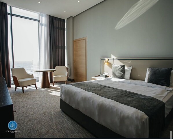 A hotel room with a large bed, two armchairs, a round table, and a view of the ocean.