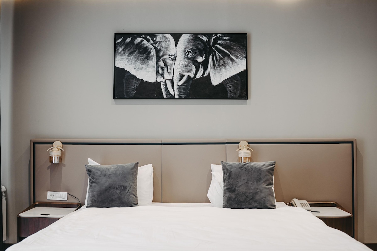 A modern hotel room with a large bed, two bedside tables, a nightstand, a wall-mounted artwork of elephants, and a ceiling light.