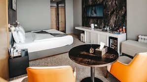 A modern hotel room with a bed, a seating area with two orange chairs, a round table, a minibar, and a TV mounted on the wall. - Port Marine Hotel By HOLLYWOOD (Baku)