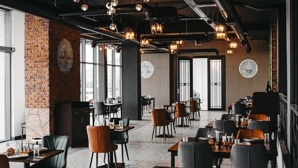 A restaurant with a modern industrial design, featuring exposed ductwork, brick walls, and a mix of round and rectangular tables. - Port Marine Hotel By HOLLYWOOD (Baku)