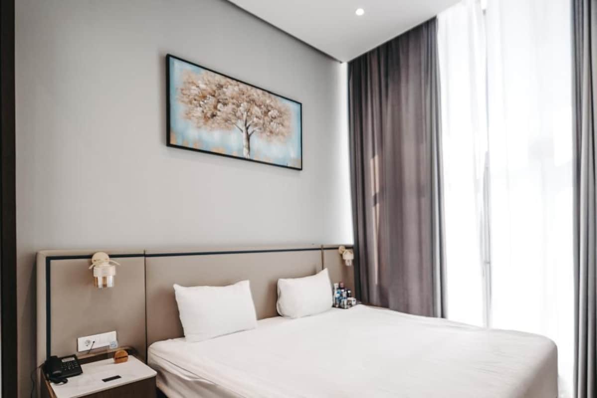 Une chambre d’hôtel moderne avec un grand lit, une table de chevet avec un téléphone et une lampe, un tableau au mur et une vue sur l’extérieur par une fenêtre.