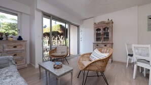 A spacious room with a large sliding glass door leading to a garden, a wicker chair with a cushion, a wooden cabinet, and a white chair.