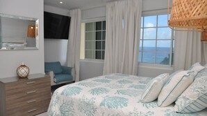 Een slaapkamer met een bed, een stoel, een dressoir, een televisie en een raam met uitzicht op zee.