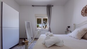 Un dormitorio con cama, armario, ventana con cortinas y una mesita de noche con lámpara.