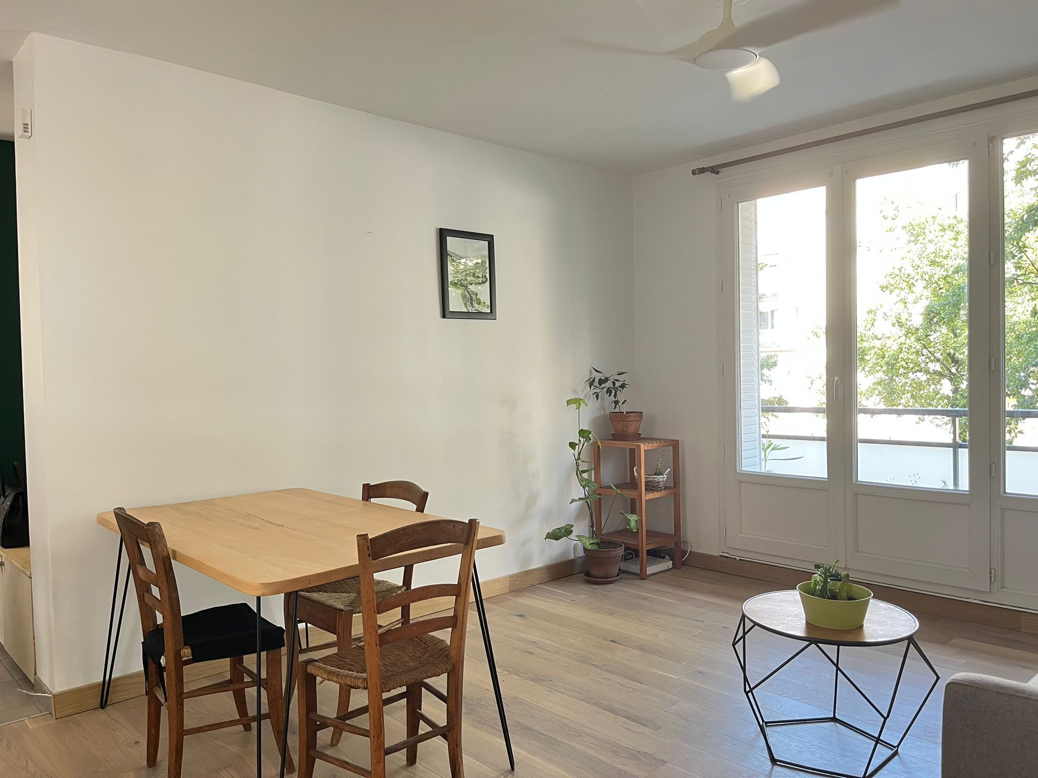 Ein Zimmer mit einem Holzesstisch und Stühlen, einem kleinen runden Tisch mit einer Pflanze und einem Fenster mit Blick auf Grün.