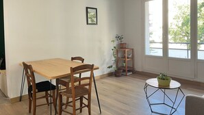 Une pièce comprenant une table à manger et des chaises en bois, une petite table ronde avec une plante, et une fenêtre donnant sur un paysage verdoyant.