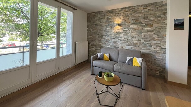 Un salon moderne avec un mur en pierre, un canapé gris agrémenté de coussins jaunes, une table basse ronde en bois et de grandes fenêtres donnant sur des arbres et un parking.