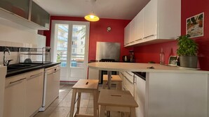 Une cuisine compacte avec des meubles blancs, un tabouret en bois et une vue sur un balcon extérieur, accessible par une porte vitrée.