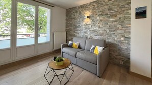 Un salon moderne avec un mur en pierre, un canapé gris agrémenté de coussins, une table basse ronde en bois sur laquelle est posée une plante, et de grandes fenêtres offrant une vue sur des arbres et un bâtiment à l’extérieur.