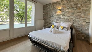 Une chambre avec un mur en pierre, un lit avec une literie blanche et une vue sur un arbre à travers de grandes fenêtres.