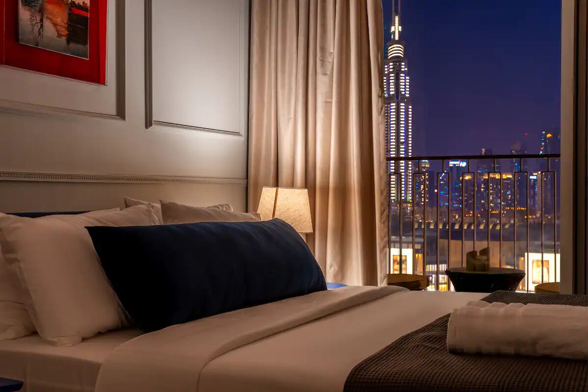 Habitación de hotel con una cama, una mesita de noche, una lámpara y vista a un paisaje urbano nocturno.