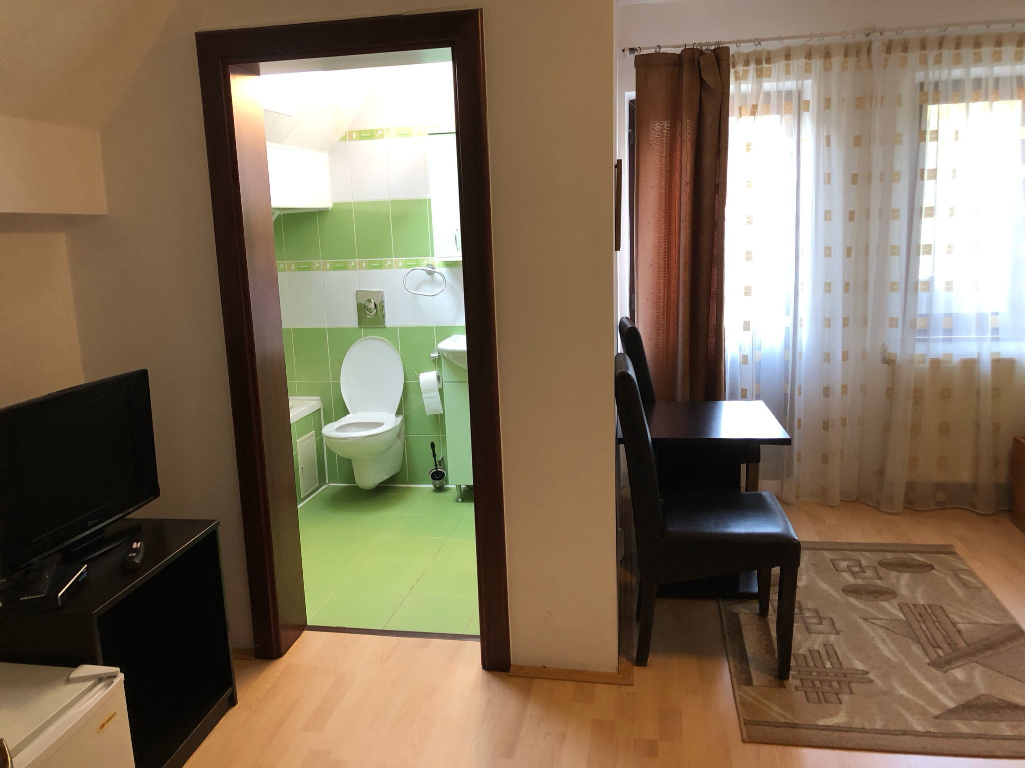A room with a television, a dining table with chairs, a rug, and a view of a bathroom with a toilet and sink.