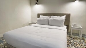 Een net opgemaakt bed met witte lakens en kussens, een nachtkastje met een lamp, en een vloer met een patroon.