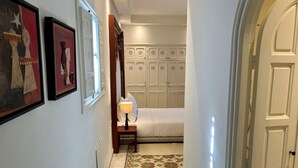 A narrow hallway with a bed, a nightstand, and a wall-mounted lamp. There are framed artworks on the wall and a door with a decorative panel. - Chambre Rabaa   Dar Kbira (Tunis)