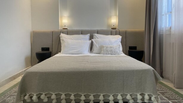 A neatly made bed with a grey bedspread and white pillows, flanked by two bedside tables with lamps, in a room with a patterned carpet and a large window with curtains.