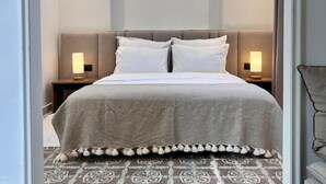 A neatly made bed with a grey duvet, two white pillows, and a decorative fringe at the foot of the bed. There are two bedside lamps on wooden nightstands. The room has patterned floor tiles and a grey carpet.