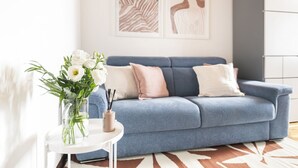 Un salon moderne avec un canapé bleu, des coussins blancs et roses, un vase en verre contenant des fleurs blanches et une petite table sur laquelle est posé un chandelier.
