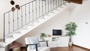 Un escalier avec une rampe en métal, un canapé blanc avec des coussins, un téléviseur posé sur un meuble blanc et deux plantes en pot.