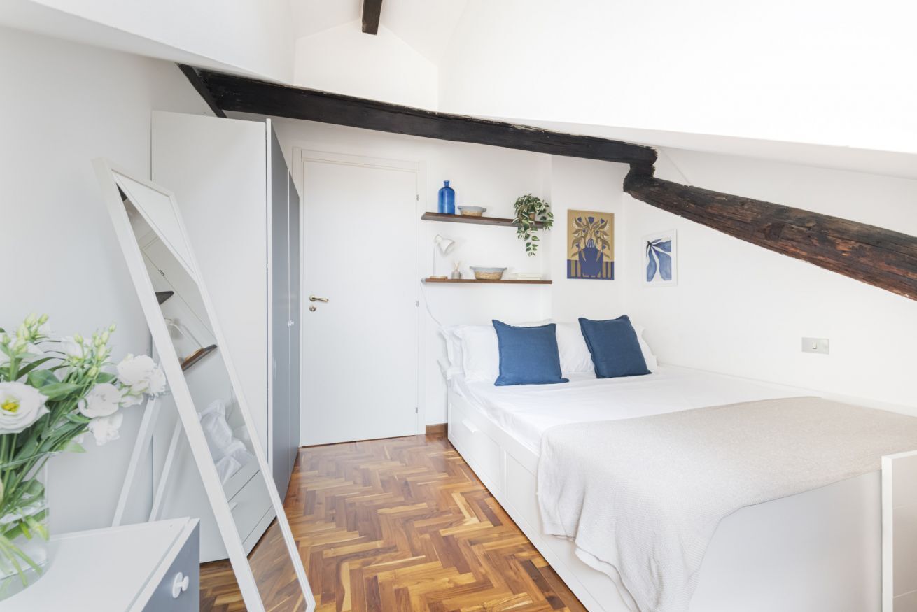 Ein Schlafzimmer mit einem Bett, weißen Wänden, Holzboden, einem weißen Schminktisch, einem Spiegel und einer Pflanze auf einem Regal.