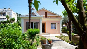 Una casa de dos pisos con fachada rosa, una entrada principal y un pequeño jardín con plantas en macetas y un banco de piedra.