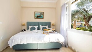 Una cama bien hecha con cabecera azul, un cuadro de barcos en la pared, y una ventana que da a un árbol y edificios.