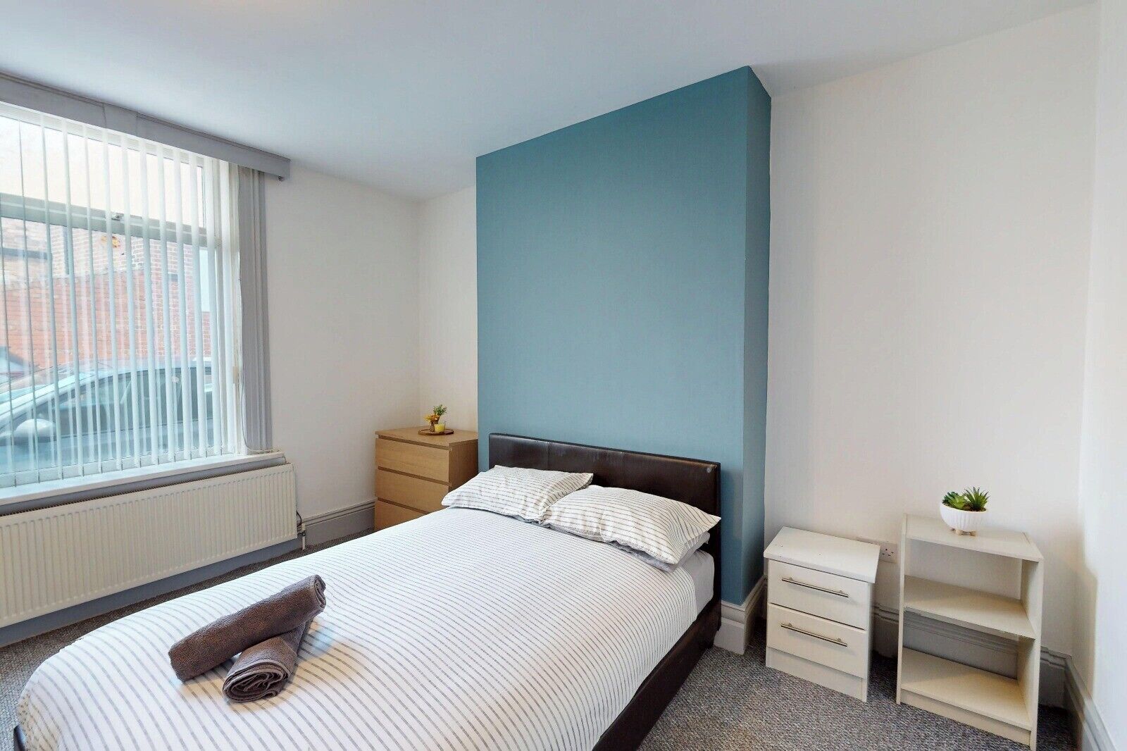 Ein Schlafzimmer mit einem Bett, einem Nachttisch, einer Kommode, einem Heizkörper und einem Fenster mit Jalousien.