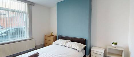 Ein Schlafzimmer mit einem Bett, einem Nachttisch, einer Kommode, einem Heizkörper und einem Fenster mit Jalousien.