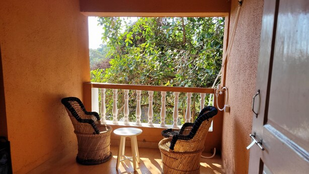A balcony with wicker chairs and a small table, a white railing, and a view of greenery outside. - Charming 1-bedroom apartment with WiFi, AC in vibrant Goa (Goa)
