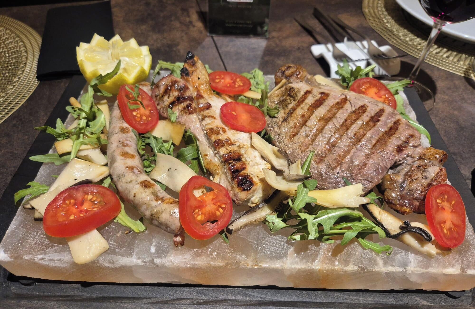 A meal with grilled meat, sausages, and vegetables on a slate plate.