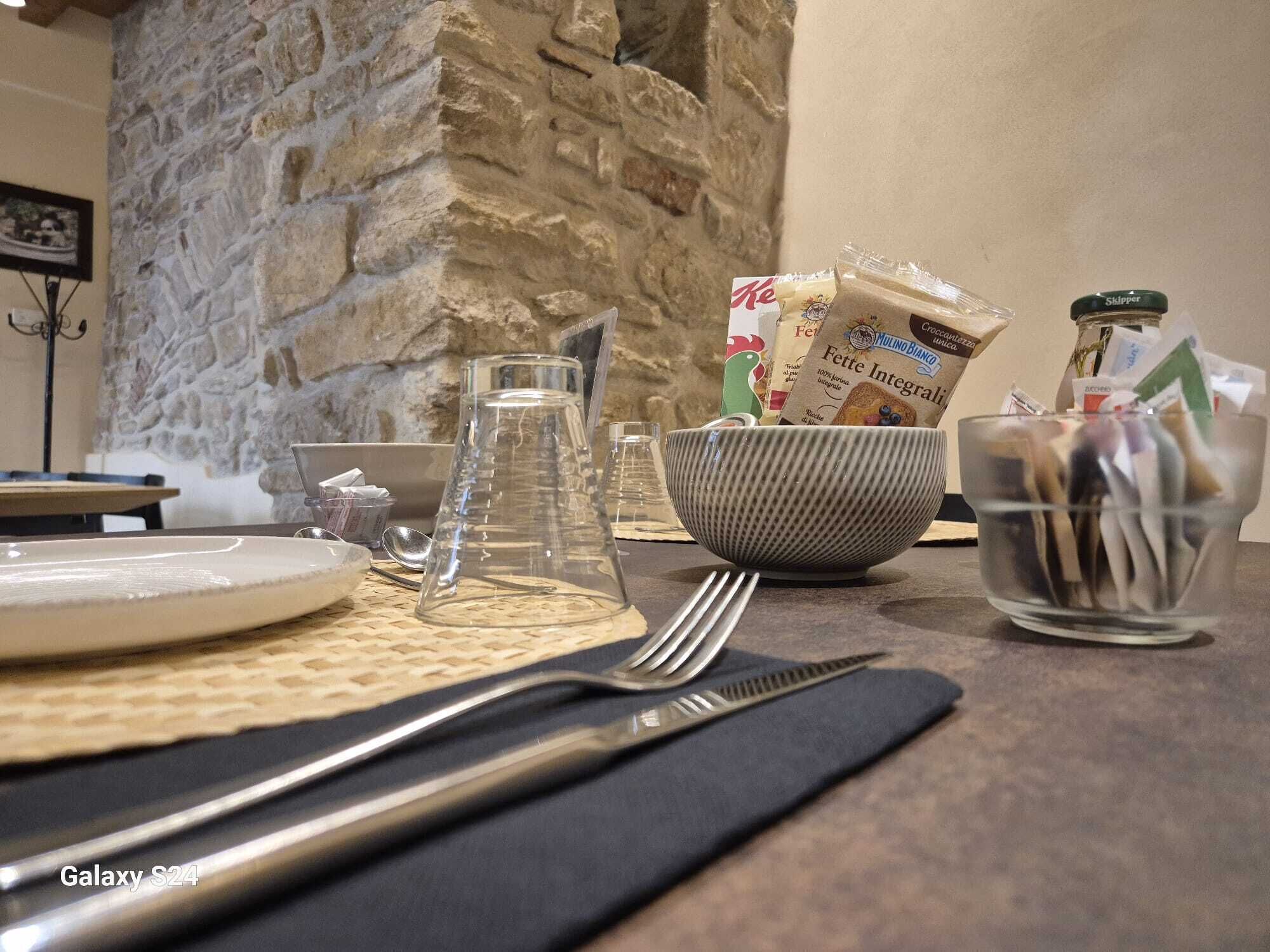 A dining table with a stone wall, a bowl of utensils, a glass pitcher, and a bag of Fette Integrali pasta.