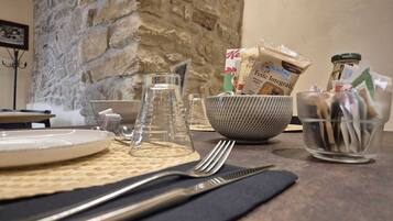 A dining table with a stone wall, a bowl of utensils, a glass pitcher, and a bag of Fette Integrali pasta.