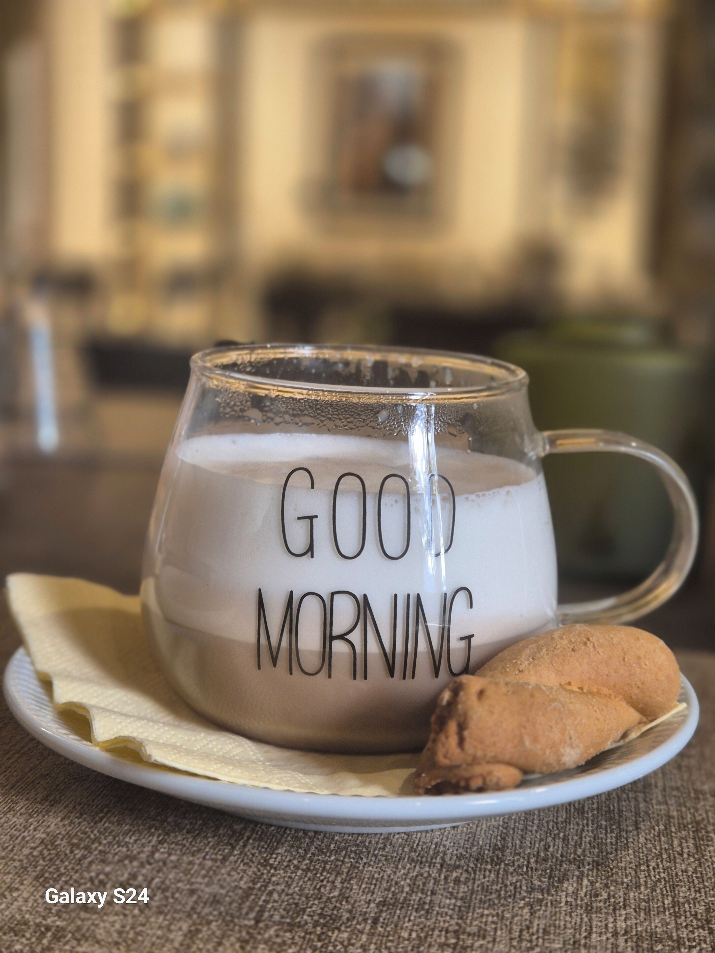 A clear glass mug with "GOOD MORNING" written on it, placed on a plate with a cookie beside it.