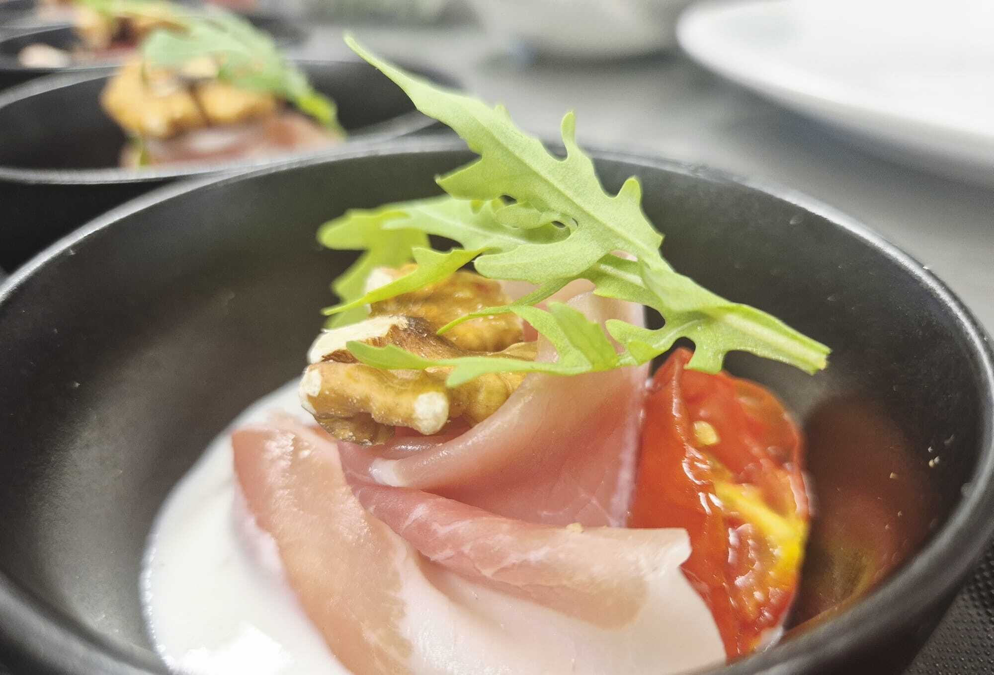 A dish with prosciutto, tomato, and greens in a black bowl.